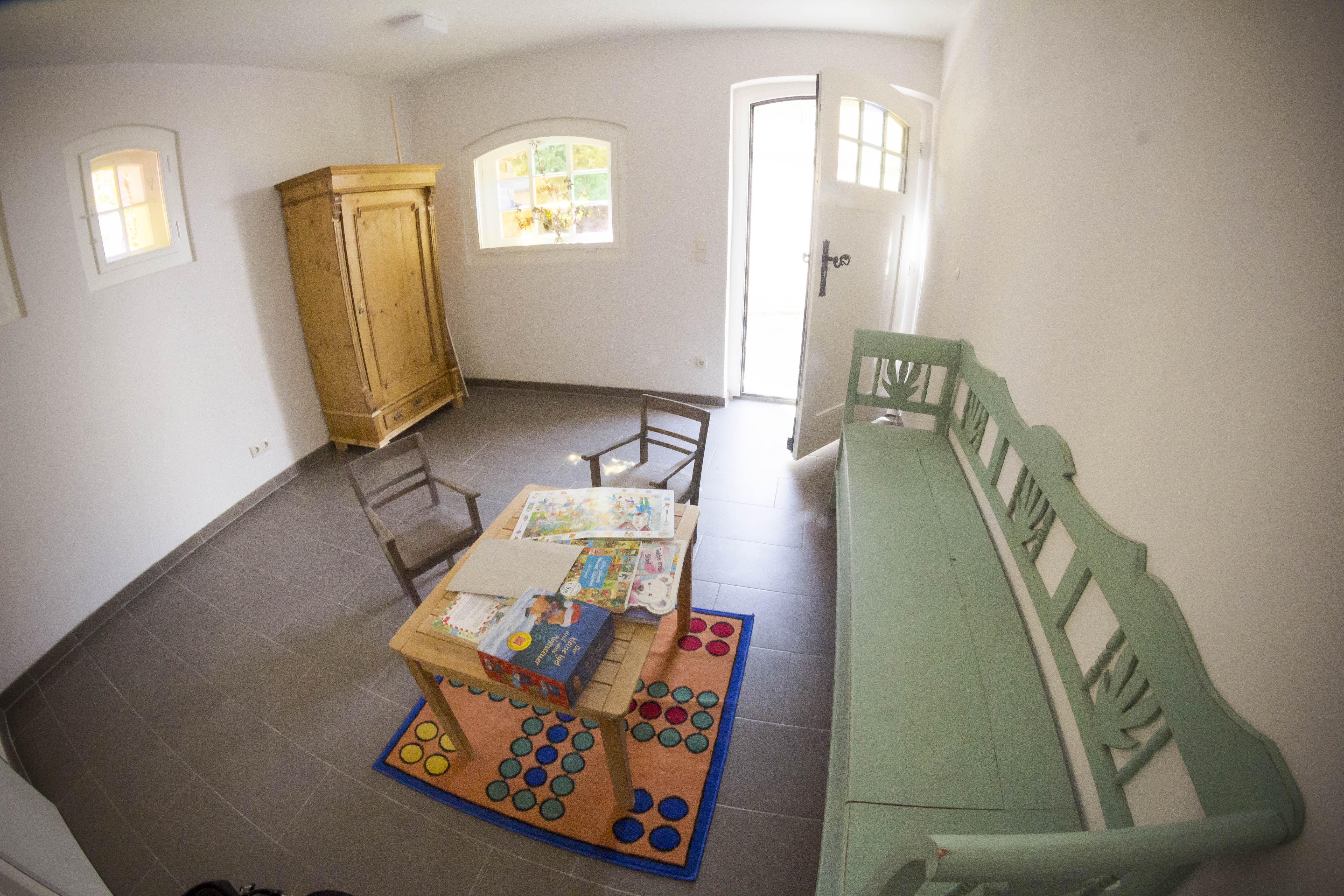 Kinderbetreuungsraum mit einer grünen Bank und einem gelben Schrank. In der Mitte des Raumes befindet sich ein Teppich mit Mensch ärger Dich nicht und darauf ein kleiner Tisch und zwei kleine Stühle.